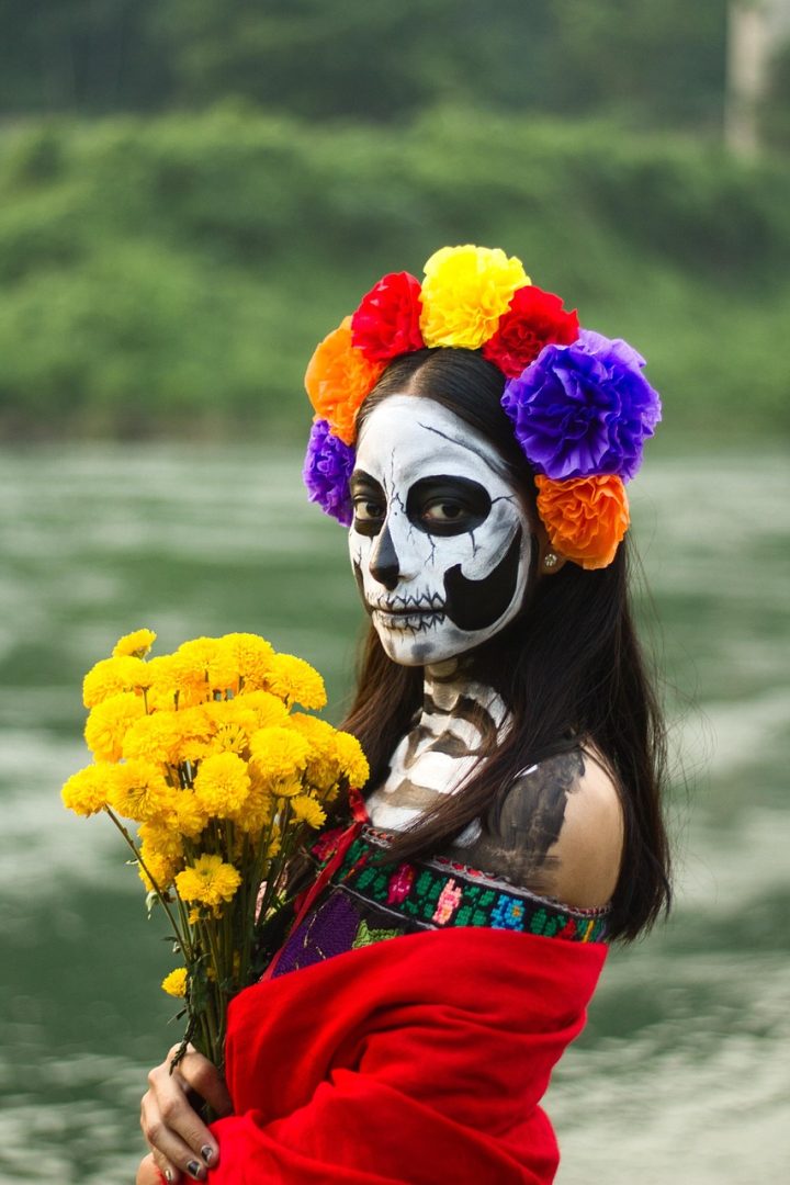 Mujer con maquillaje de catrina para el Día de Muertos en México, sosteniendo flores de cempasúchil y usando una corona de flores de colores vivos (rojo, amarillo, morado y naranja). La fotografía resalta la tradición cultural mexicana que honra a los difuntos, mostrando la conexión entre el arte, la identidad y el simbolismo de esta festividad. Imagen ideal para ilustrar artículos sobre Día de Muertos, cultura mexicana, catrinas y tradiciones ancestrales.